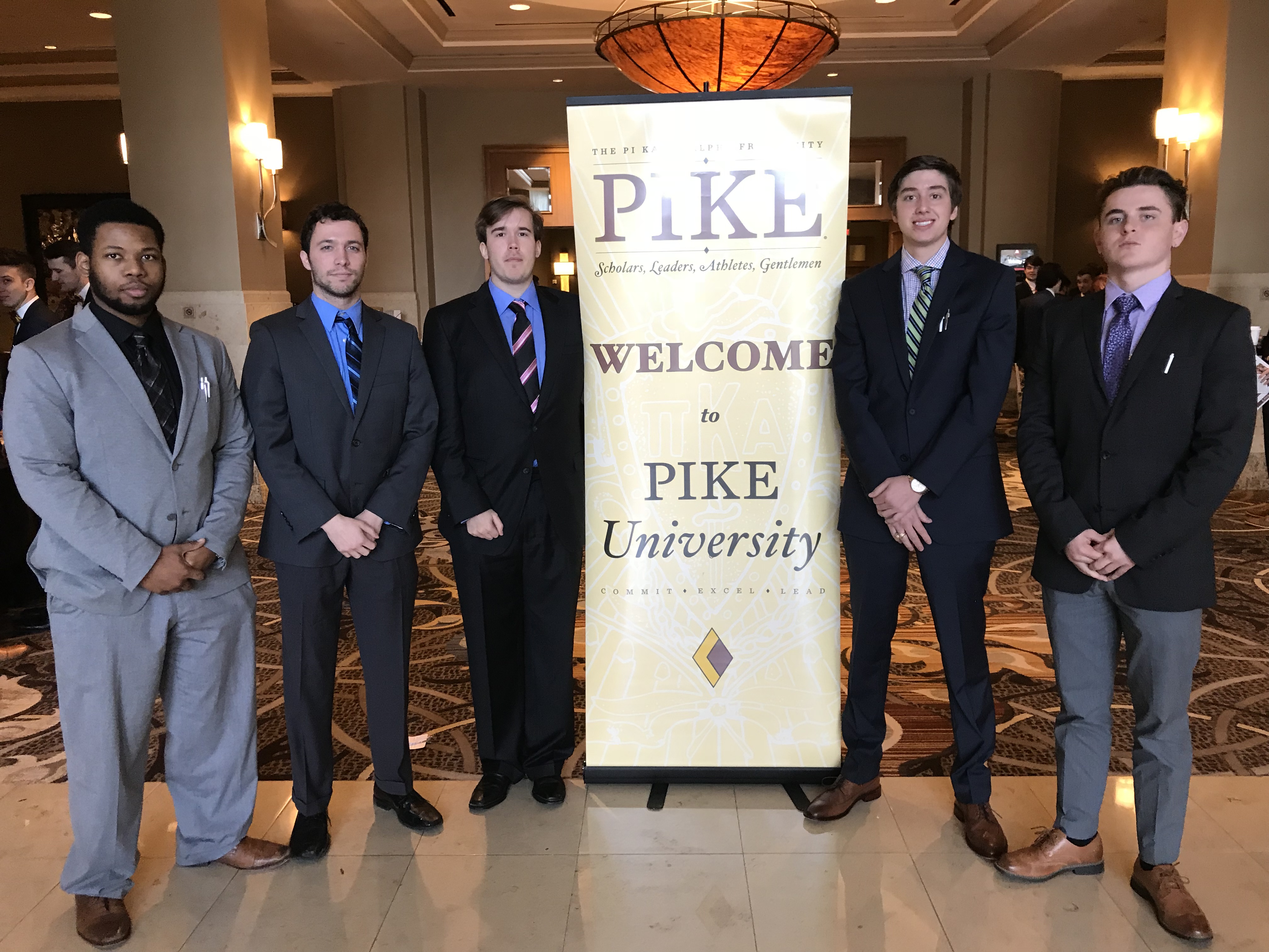 5 men in suites standing next to a sign that welcomes them to Pike University.